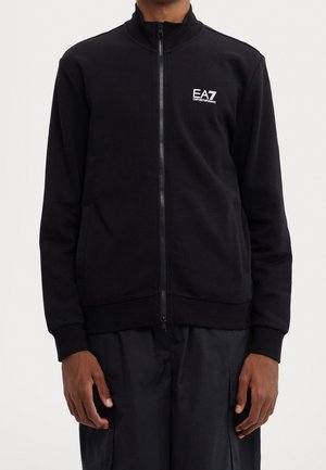 Person trägt eine schwarze Jacke mit Reißverschluss und EA7 Emporio Armani Logo auf der Brust sowie schwarze Hose und steht vor einem schlichten hellen Hintergrund.