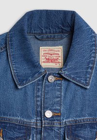 Denimjakke i middels blå med spiss krage, sølvknapper og oransje søm. Har en fremtredende merkelapp på innsiden.