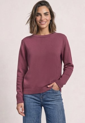 Femme souriante portant un pull en maille mauve à manches longues et un jean bleu, une main dans la poche, sur un fond uni pâle.