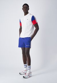 Polo bianco con maniche rosse e blu, abbinato a pantaloni corti blu con loghi. Il modello indossa sneakers bianche e calzini a righe.