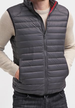 Mężczyzna ubrany w zapinaną na zamek ciemnoszarą pikowaną kamizelkę na beżowym swetrze, z rękami w kieszeniach kamizelki.