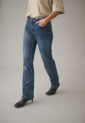 Person iført blå straight-leg jeans og sorte støvler med hæl, med den ene hånd i lommen og beige top synlig.