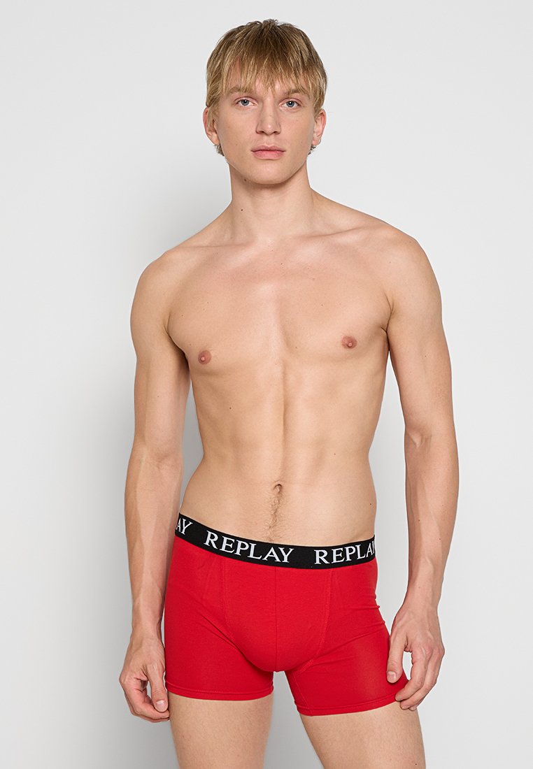 Männliches Model mit kurzem blonden Haar, das rote Boxershorts mit schwarzem Bund trägt, auf dem "REPLAY" steht, vor einem schlichten hellen Hintergrund.