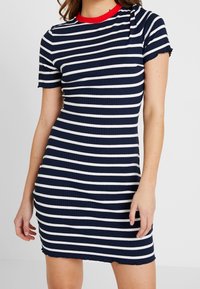 Granatowa i biała sukienka w paski o prążkowanej fakturze, z krótkimi rękawami i okrągłym czerwonym dekoltem. Charakteryzuje się dopasowanym krojem z postrzępionym dołem.