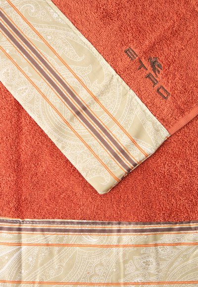 Serviette orange avec bandes de tissu beige à motif et logo de la marque brodé en fil marron foncé sur une surface texturée.