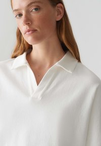 Weißes Poloshirt mit spitzem Kragen und leicht strukturierter Oberfläche, das eine lockere Passform und ein tiefes V-Ausschnitt-Design aufweist.