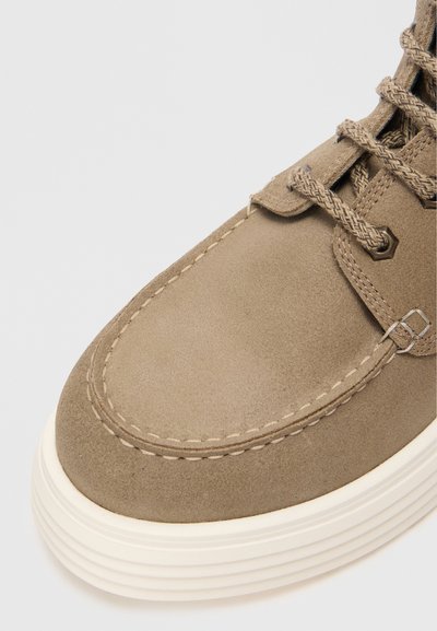 Zapato de ante beige con punta redondeada, superficie texturizada, suela de goma blanca contrastante y cordones marrones con un ojal hexagonal.