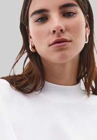 Bílá mikina s vysokým límcem a hladkou strukturou. Modelka má zlaté kruhové náušnice; vlasy jsou hnědé a mírně vlnité.