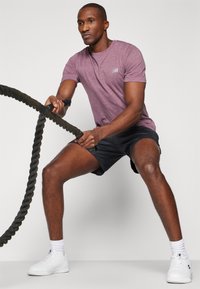 Homme athlétique en chemise à manches courtes violette et short noir, tenant une corde de combat, portant des baskets blanches et des chaussettes de sport sur fond uni.