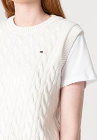 Vit kabelstickad väst med ribbade kanter, prydd med en liten logotyp på bröstet, bärs över en vit kortärmad skjorta.