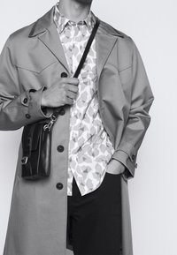 Beige trenssitakki, jossa on kaksiriviset napit, kuviollinen vaalea paita alla ja pieni musta olkalaukku.