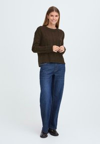 Ciemnozielony sweter z dzianiny z wzorem w warkocz, z ściągaczami przy mankietach i na dole. Zestawiony z prostymi niebieskimi dżinsami i czarnymi butami.