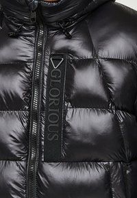 Zwarte pufferjas met een glanzende afwerking, horizontaal doorgestikt patroon, rits aan de voorkant en een opvallende logoband op de borst.