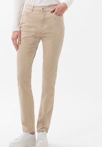 Pantalones slim-fit beige hechos de una mezcla de algodón, con bolsillos frontales, cremallera y una textura suave, combinados con zapatillas blancas.