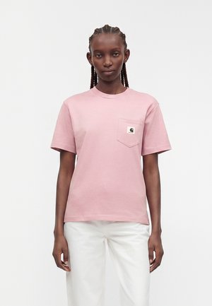 Kobieta z zaplecionymi włosami ubrana w jasnoróżowy T-shirt z krótkim rękawem i kieszonką na piersi oraz białe spodnie, stojąca na jednolitym jasnym tle.
