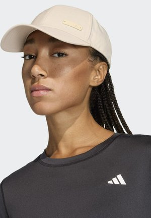 Gorra beige clara con visera curva, con una etiqueta rectangular, acompañada de una camiseta negra ajustada que muestra un logo blanco en el pecho.