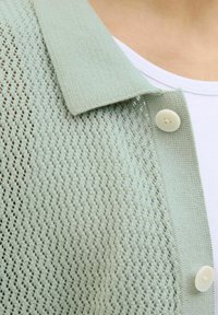Svijetlo zelena pletena cardigan jakna s ovratnikom, koja se odlikuje labavim tkanjem i jednim zatvaračem na bijeli gumb.