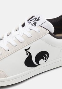 Baskets blanches avec un dessus en daim et cuir, présentant un design de logo noir et des lacets blancs. Des accents noirs sur le talon apportent un contraste.