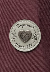Leren label op een bordeauxkleurige stof. Het label heeft een hartvormig zilveren embleem, decoratieve stiksels en de tekst "Ragwear since 1997".