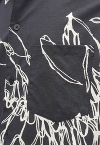 Camisa preta com botões e bolso no peito, apresentando arte abstrata em linhas brancas que lembram girafas e plantas.