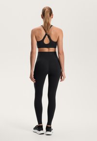 Czarny sportowy stanik z krzyżującymi się ramiączkami i otworem w kształcie klucza z tyłu, w zestawie z czarnymi legginsami o wysokim stanie i czarnymi butami sportowymi.