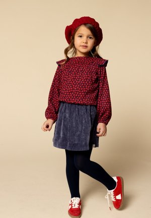 Rode bloempatronde lange mouw top, met ruffles op de schouders, marineblauwe corduroy rok, marineblauwe maillots, en rode sneakers met witte accenten.