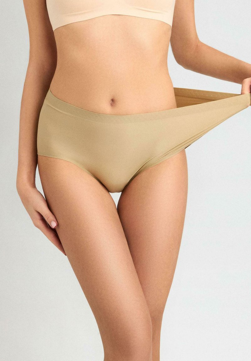 Beżowa, bezszwowa bielizna w stylu bikini o gładkiej fakturze i szerokim, elastycznym pasie, lekko rozciągająca się podczas trzymania w dłoni.
