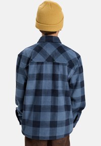 Camicia flanella a quadri blu e navy con maniche lunghe, orlo curvo e dettagli sulla schiena, abbinata a un berretto di maglia giallo.