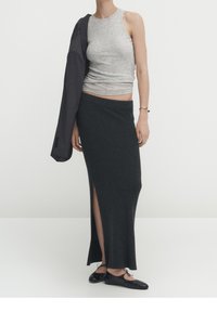 Top grigio senza maniche, gonna di maglia grigio scuro con spacco laterale, ballerine nere e una giacca scura appoggiata sull'avambraccio.