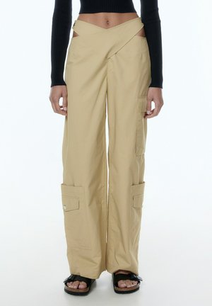 Beige wide-leg cargo pants with side cutouts, featuring two side pockets and a smooth texture. Paired with chunky black sandals.