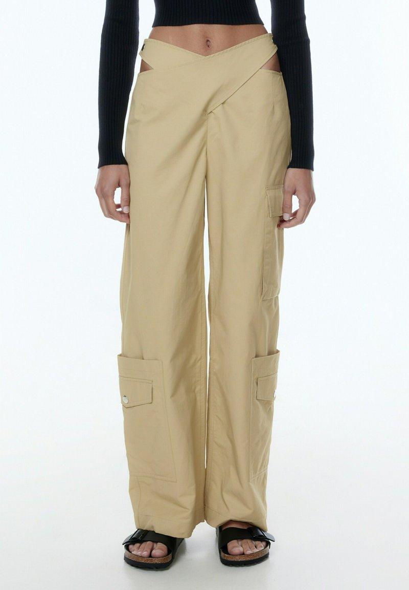 Beige wijd uitgesneden cargobroek met zij-inzendingen, voorzien van twee zijzakken en een gladde textuur. Gepaard met chunky zwarte sandalen.
