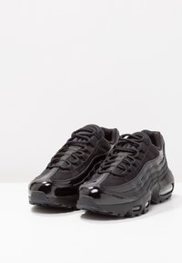 Svarta sportskor med en texturerad mesh och ovandel i lackläder. Har en böjd design, tjock sula och synlig luftkuddning.