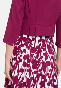 Dopasowana, krótką magentową kurtkę z rękawami do trzech ćwiartek, zestawioną z białą spódnicą z wyrazistym, bordowym kwiatowym nadrukiem i plisami.