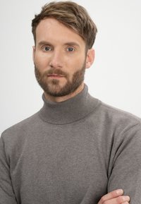 Grauer Rollkragenpullover aus weichem Material, mit geripptem Ausschnitt und nahtlosem Design. Modell mit kurzem Haar und Bart.