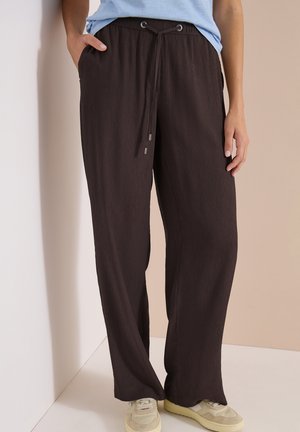 Personne porte un pantalon ample marron foncé texturé avec une ceinture à cordon et des baskets beiges, debout contre un mur d’angle beige clair et blanc.