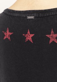 Musta t-paita, jonka takaosassa on painettu kolme punaista tähteä. Kangas tuntuu pehmeältä, siinä on lievä tekstuuri, ja paita on klassisella pyöreällä pääntiellä.