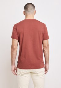 Koszulka z krótkim rękawem w rdzawej kolorystyce, wykonana z bawełny. Posiada klasyczny okrągły dekolt i prosty dół, przedstawiona z tyłu.