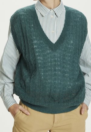 Personne portant un pull sans manches en maille bleu sarcelle par-dessus une chemise à rayures bleues avec un pantalon beige, mains dans les poches.