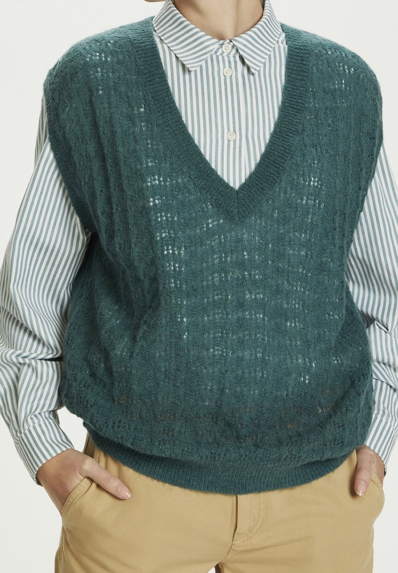 Personne portant un pull sans manches en maille bleu sarcelle par-dessus une chemise à rayures bleues avec un pantalon beige, mains dans les poches.