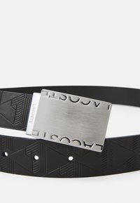 Cinturón negro de cuero texturizado con patrones geométricos, hebilla metálica plateada grabada con "LACOSTE". Superficie lisa y diseño resistente.