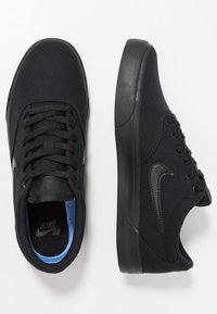 Svarta kanvas skate-skor med en platt gummisula, featuring svarta snören, en liten svart logotyp och en blå innerfoder.