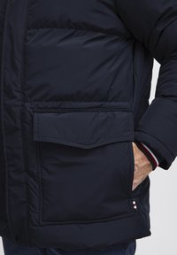 Marineblå puffjakke med lynlås, en lomme med klap og striped ærmer. Materialet virker glat og isoleret, hvilket giver varme.