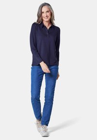 Navyblaues langärmliges Polo-Shirt mit Kragen, kombiniert mit blauen Jeans. Das Modell trägt hellfarbene Sneaker und steht vor einem weißen Hintergrund.