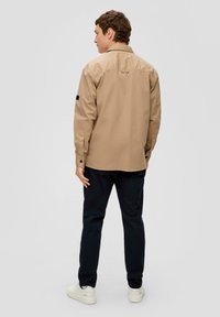 Camicia chiara marrone con bottoni e maniche lunghe, caratterizzata da una toppa nera sulla manica sinistra, abbinata a pantaloni blu navy e sneakers bianche.
