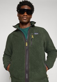 Green fleece jacket with a zippered front, high collar, textured material, and contrasting black zipper details. Features a logo on the chest.