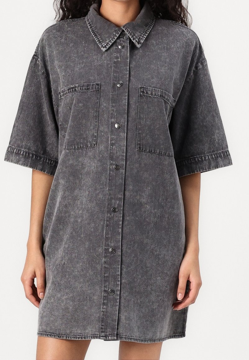 Femme portant une robe-chemise en jean noir délavé, oversize, avec de grandes poches poitrine et des manches courtes, boutonnée devant.