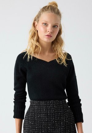 Suéter negro de punto con cuello en V y puños acanalados, combinado con una falda estampada en blanco y negro con un tejido texturizado.
