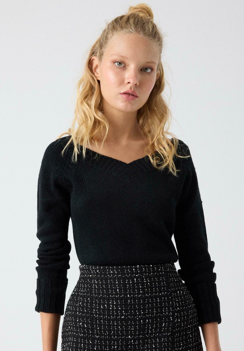 Suéter negro de punto con cuello en V y puños acanalados, combinado con una falda estampada en blanco y negro con un tejido texturizado.