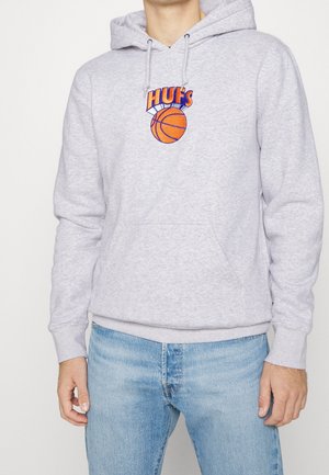 Mężczyzna w jasno szarej bluzie z kapturem z pomarańczową piłką do koszykówki i napisem "HUF'S", ubrany w jasnoniebieskie dżinsy, stojący na jednolitym tle.