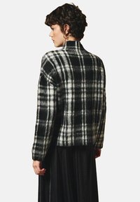 Kobieta z krótkimi kręconymi włosami nosi czarno-białą, wełnianą kurtkę w kratę z wysokim kołnierzem i długimi rękawami, zestawioną z czarną spódnicą.